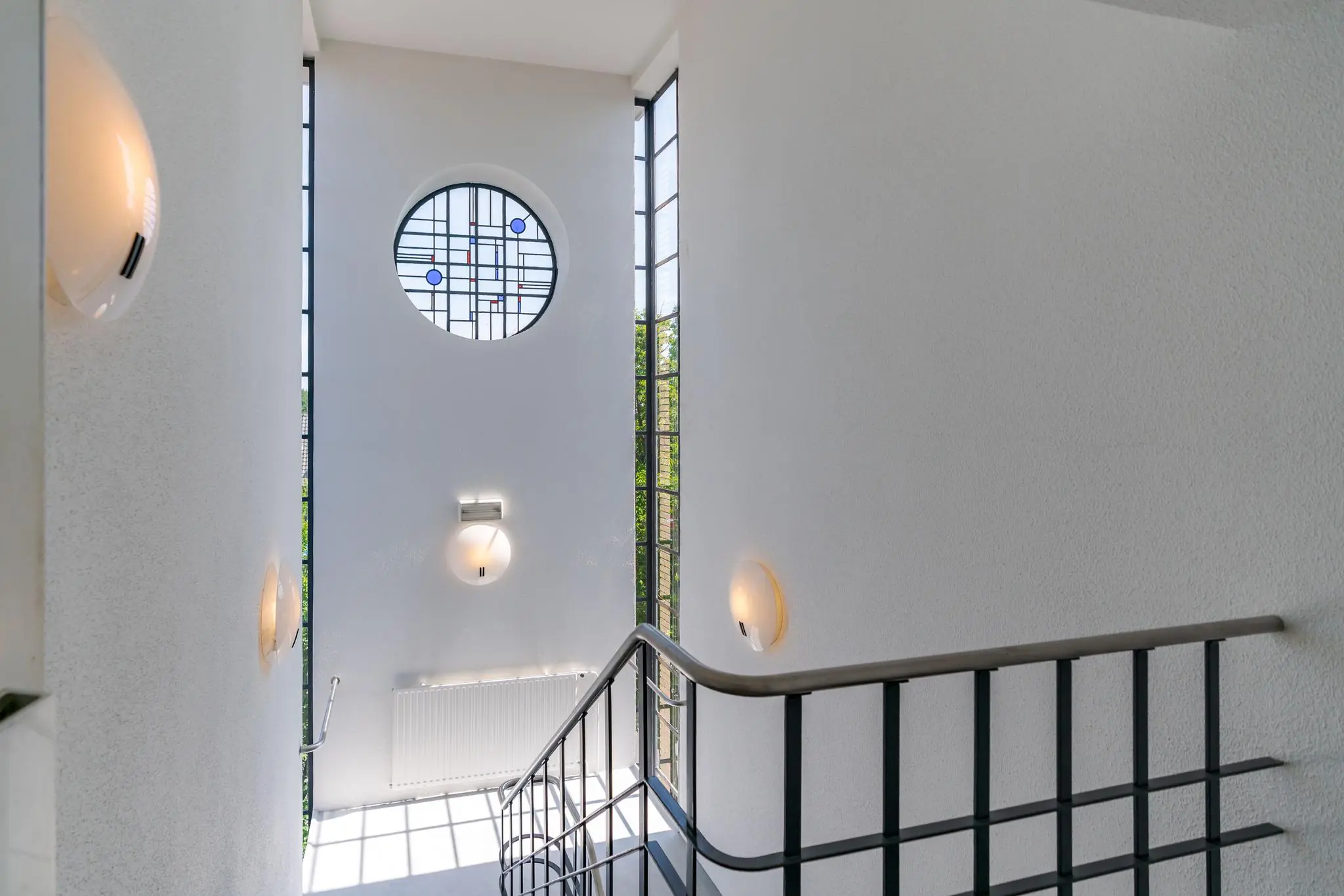 Trapportaal met ronde glas-in-loodramen en moderne wandverlichting aan de Van Alkemadelaan.